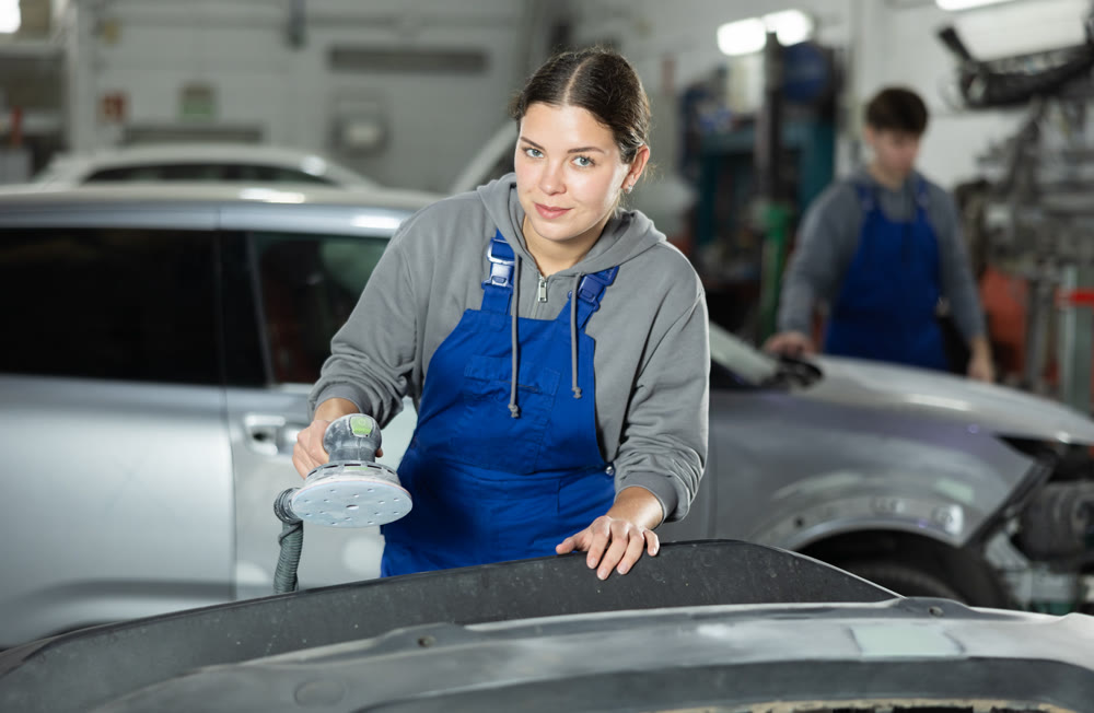 Woman auto mechanic using grinding machine prepares car parts for painting