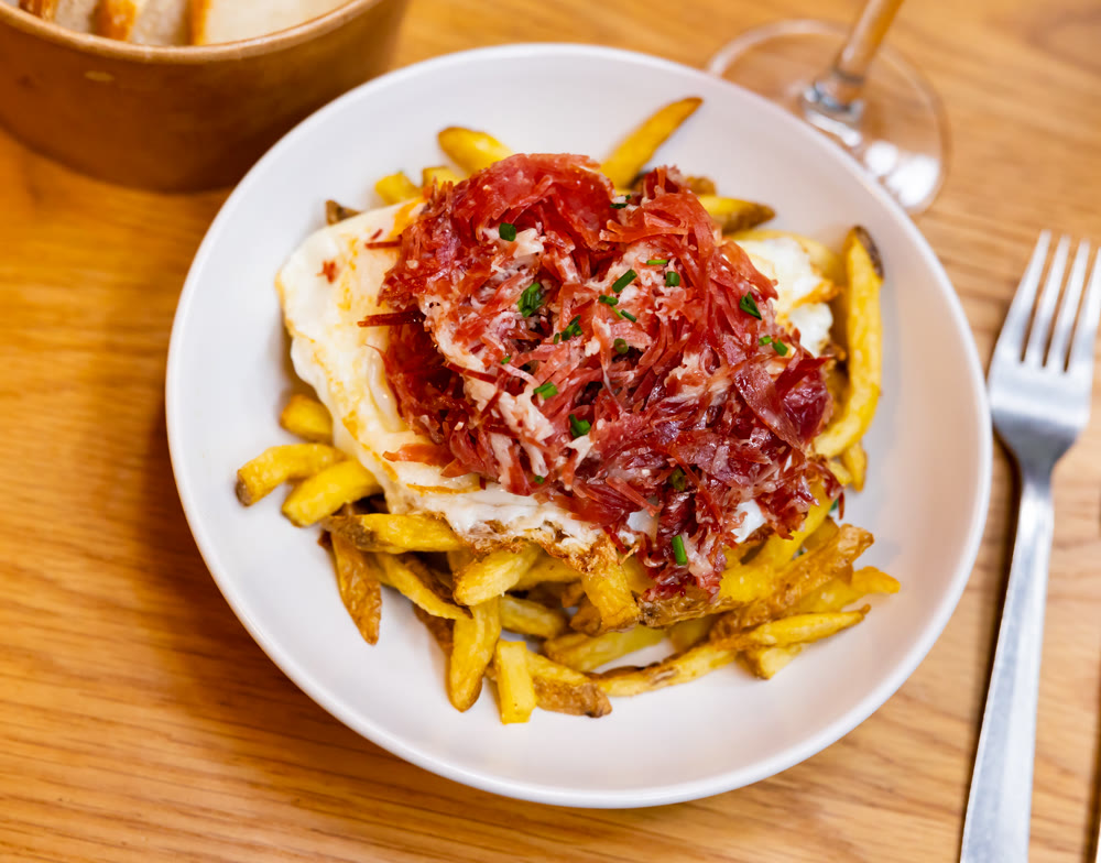 Huevos rotas served with potatoes and jamon on a plate with bread wine and cutlery